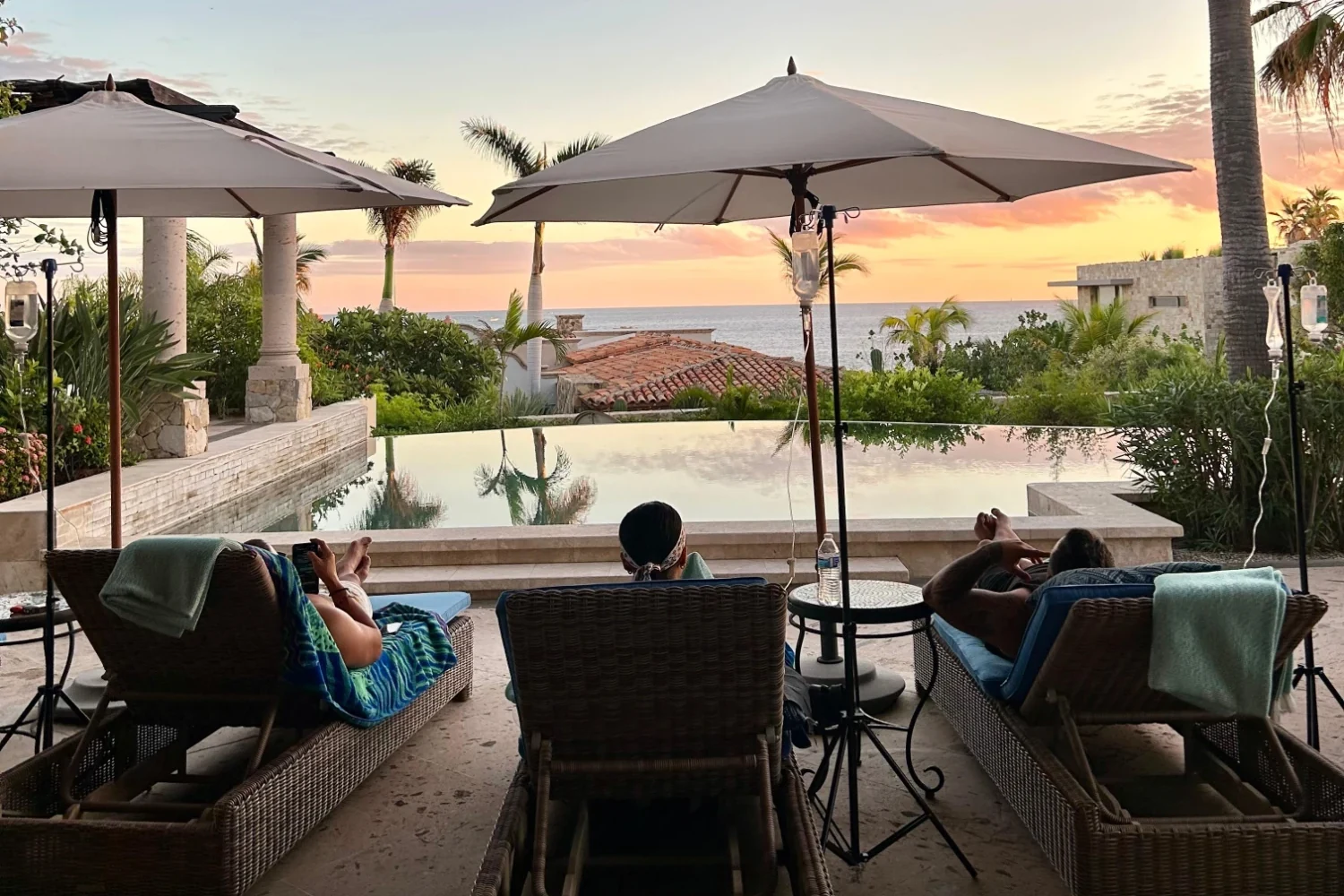 Guests receiving IV therapy on poolside loungers at a private villa in Los Cabos, with IV stands beside them and the ocean at sunset beyond the infinity pool